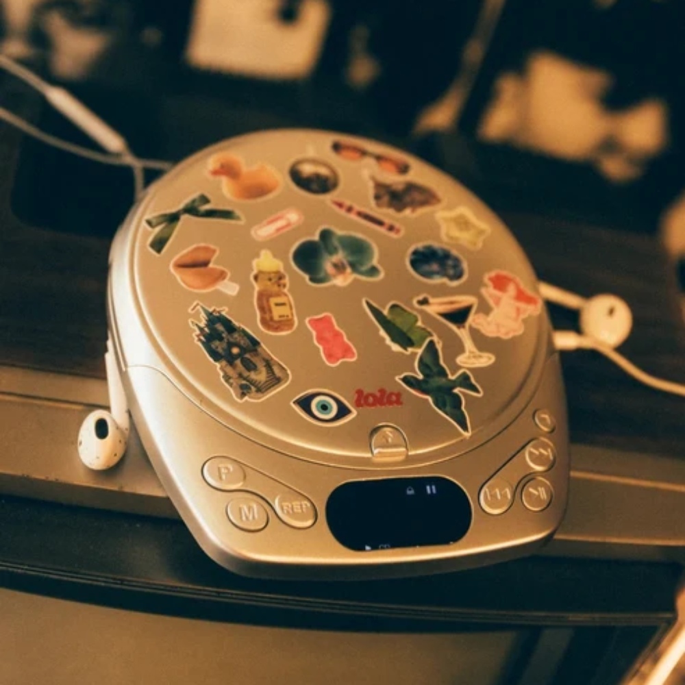 A silver portable CD player covered in colorful stickers, sitting on a dark surface with white wired earphones nearby in a dimly lit room.