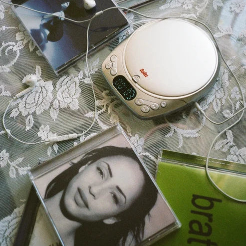 A vintage white portable cassette player with a digital display and red 'Tosca' logo, styled on a white floral lace tablecloth alongside CD cases, white earbuds, and a green book.