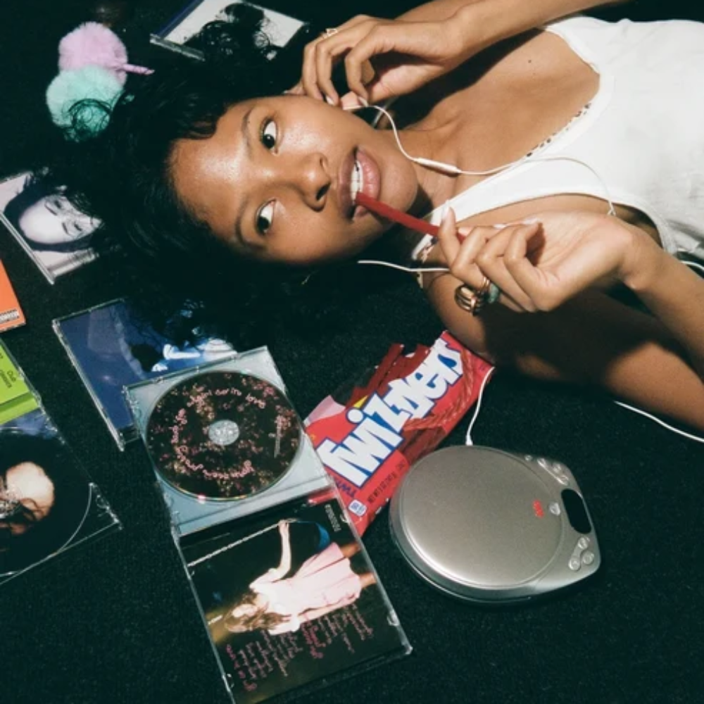 A woman lying on a dark floor surrounded by CDs, album covers, a silver CD player, a red pen, and a white tank top, with colorful pom-pom hair accessories.