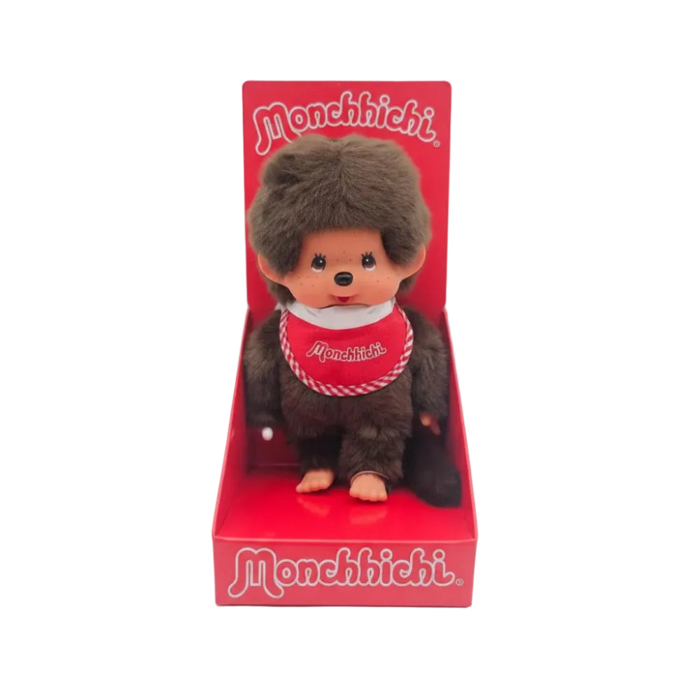 A Monchhichi plush toy sitting in its original red display box, featuring the character with brown fur, a red and white checkered bib, and the brand name 'Monchhichi' printed on the box.