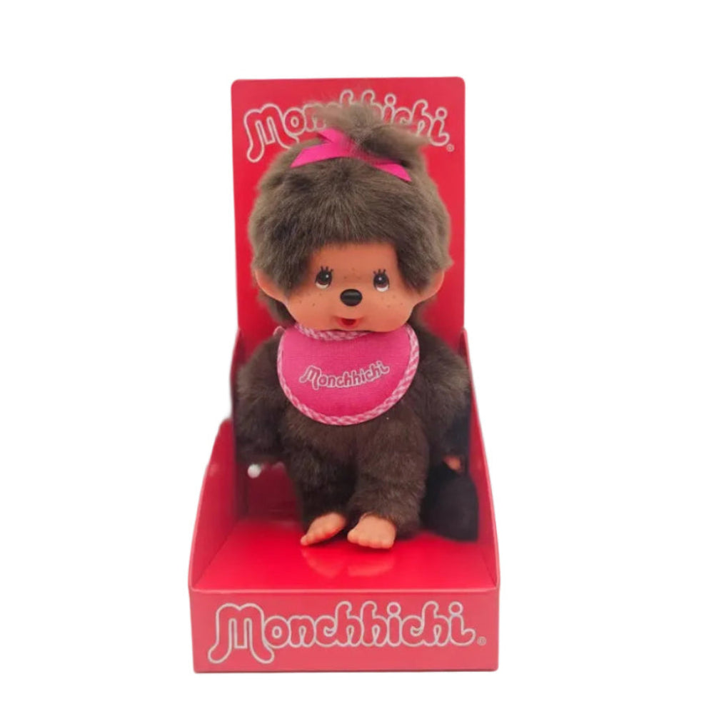 A vintage Monchichi monkey plush toy in its original red display box, featuring brown faux fur, a pink bow, and a pink bib.
