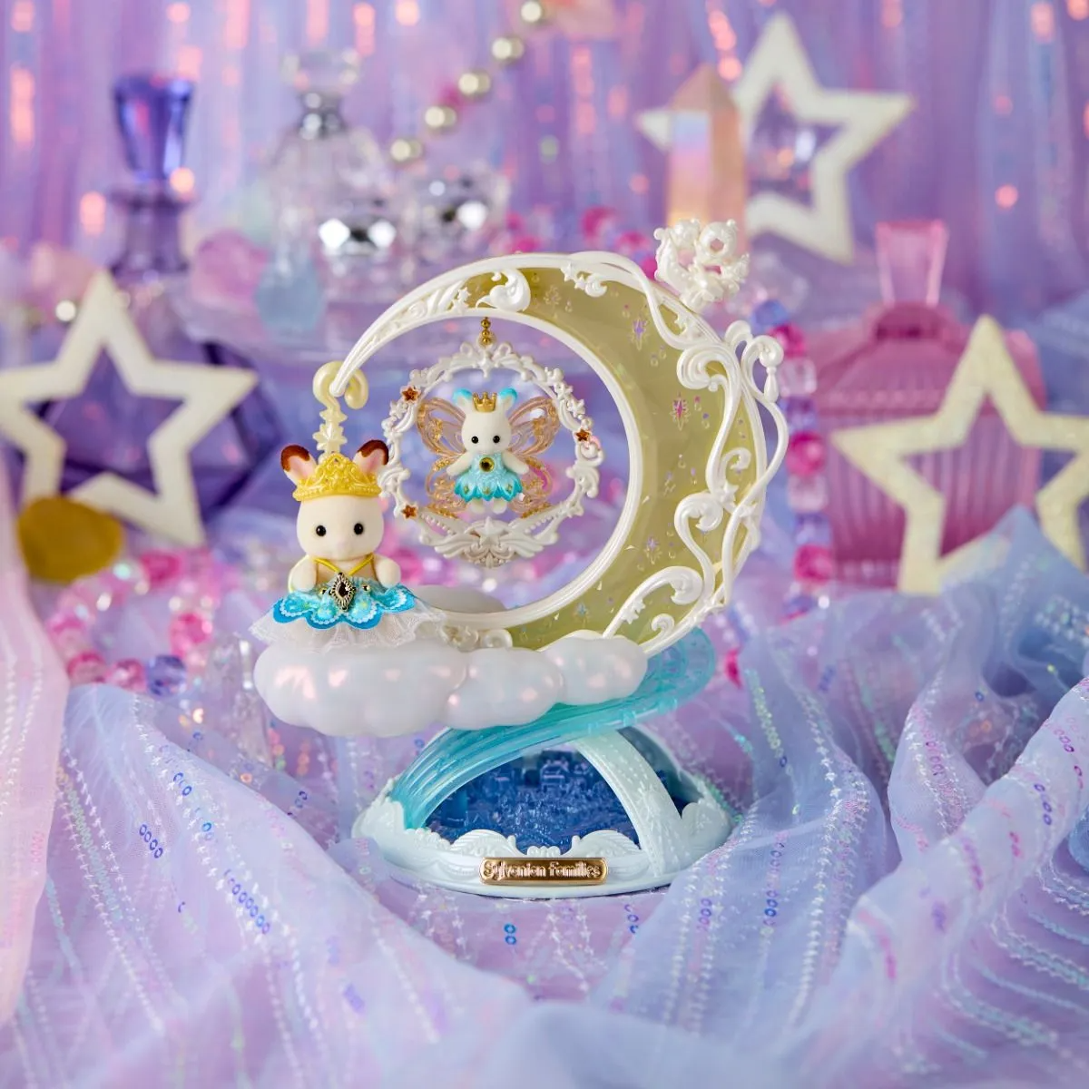 A decorative fantasy figurine or ornament featuring a small white bunny character with a gold crown and blue cape, posed beside a crescent moon frame with a fairy inside. The piece sits on a cloud-like base with a blue and white rainbow arch, surrounded by pastel pink and lavender fabric, star cutouts, and glittery decorative props.