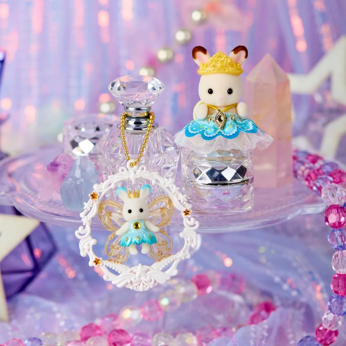 A decorative white bunny figurine with a gold crown and blue dress, displayed on a clear pedestal alongside a perfume bottle and a matching bunny ornament in a sparkling pastel setting.