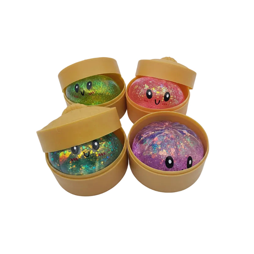 Four small squishy toys in round tan holders, each with a different glittery color and a simple face drawn on top. The colors visible are green, pink, blue, and purple, with gold glitter throughout.