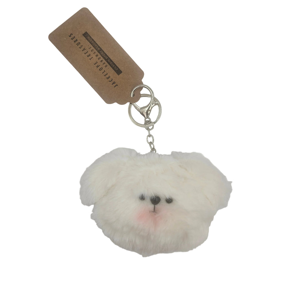 A fluffy white faux-fur keychain shaped like a small dog or bunny face, with a silver key ring and a brown paper hang tag attached, photographed on a plain light background.