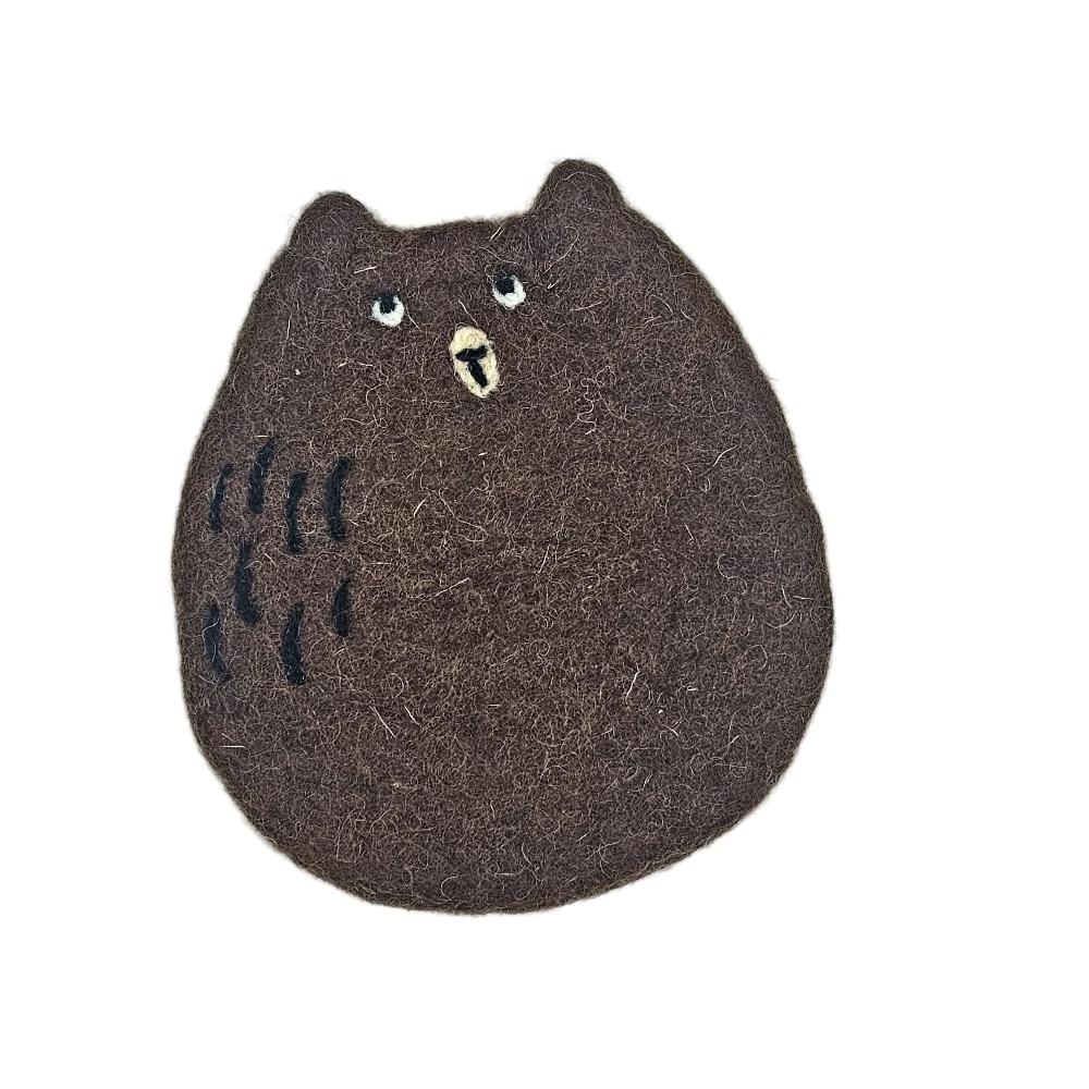 A brown felt bear-shaped coaster, likely a hair clip or pin, featuring black stitched details on the left side and a small beige beak with white eyes, set against a plain light gray background.