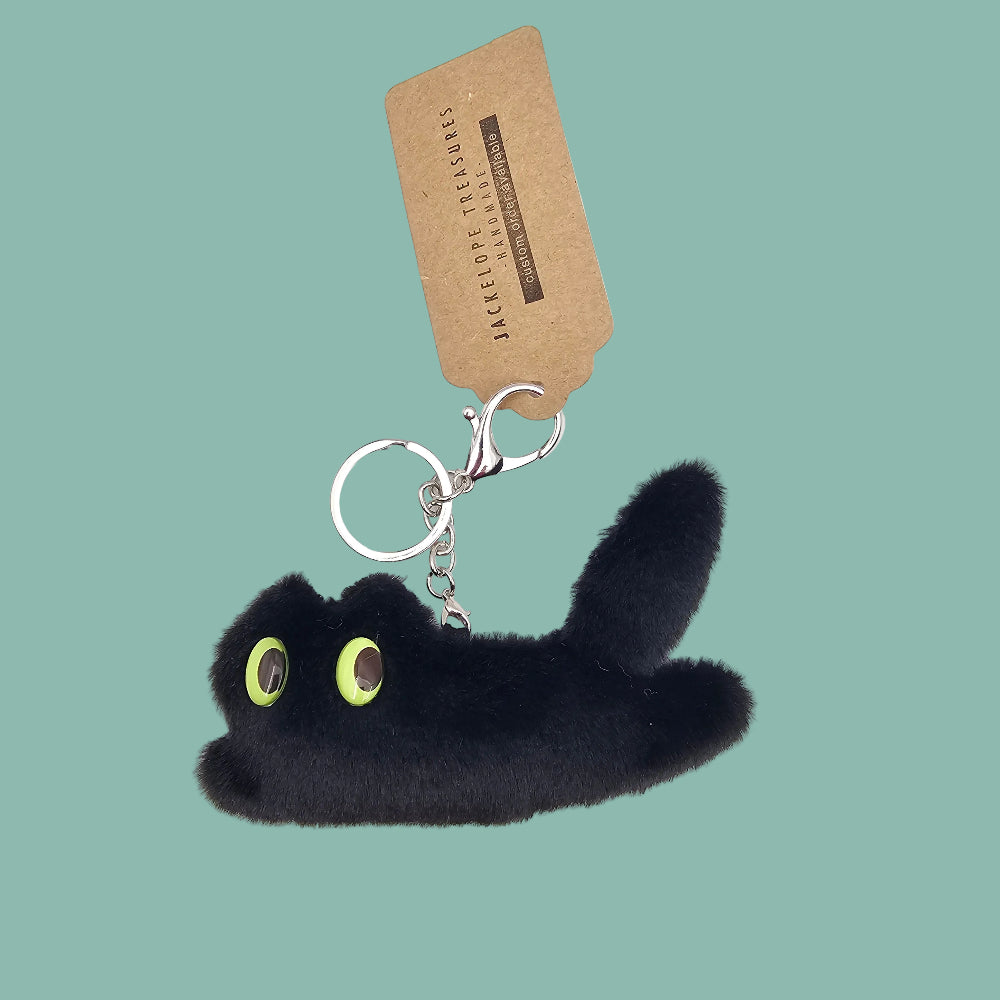 A black plush cat keychain with bright lime green eyes and a silver split ring, attached to a brown paper tag that reads 'JASPEUDE TRAZES'.