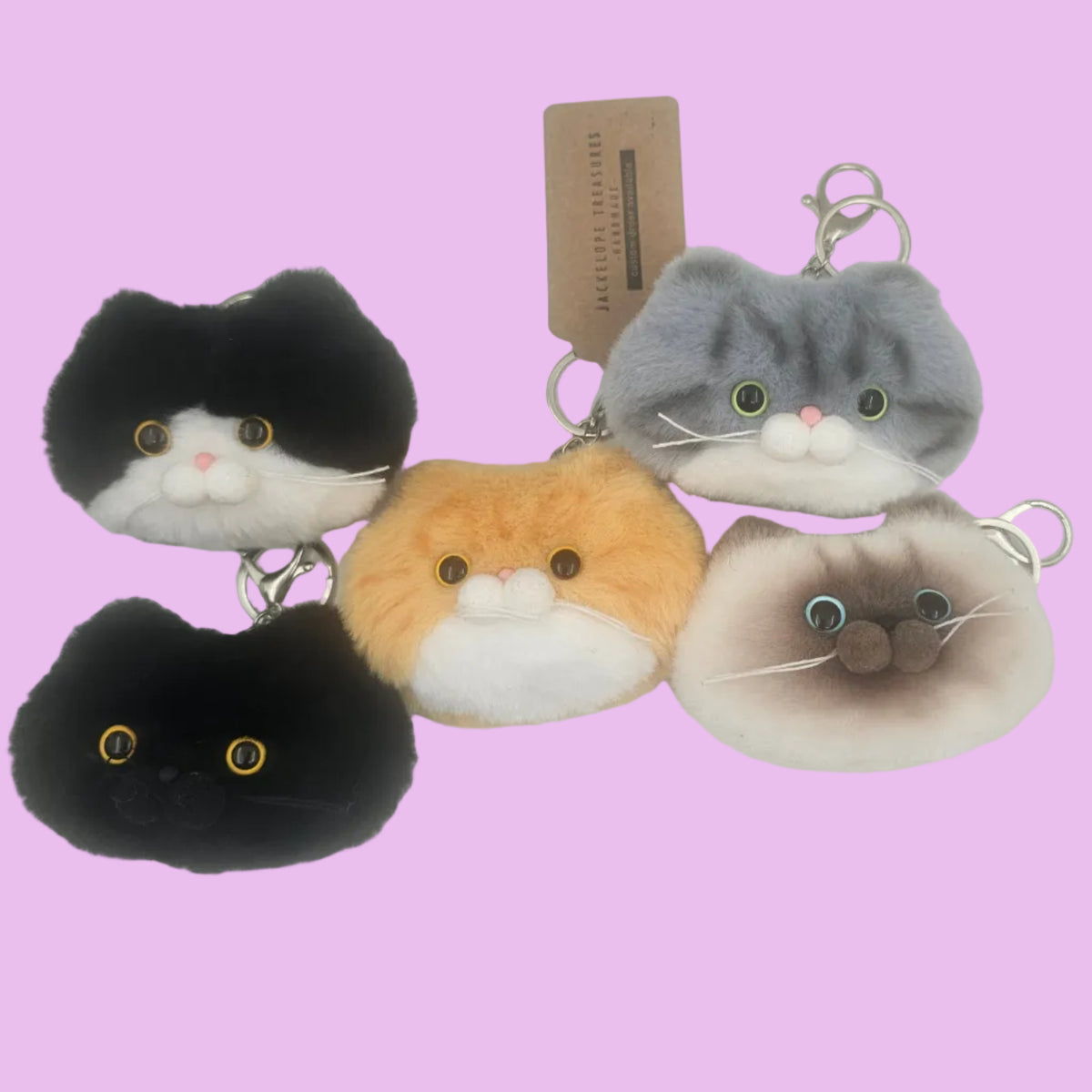 Four plush cat-shaped keychains in different colorways (black and white, orange and white, gray and white, and brown and white) arranged on a plain white surface with a small paper tag behind them.