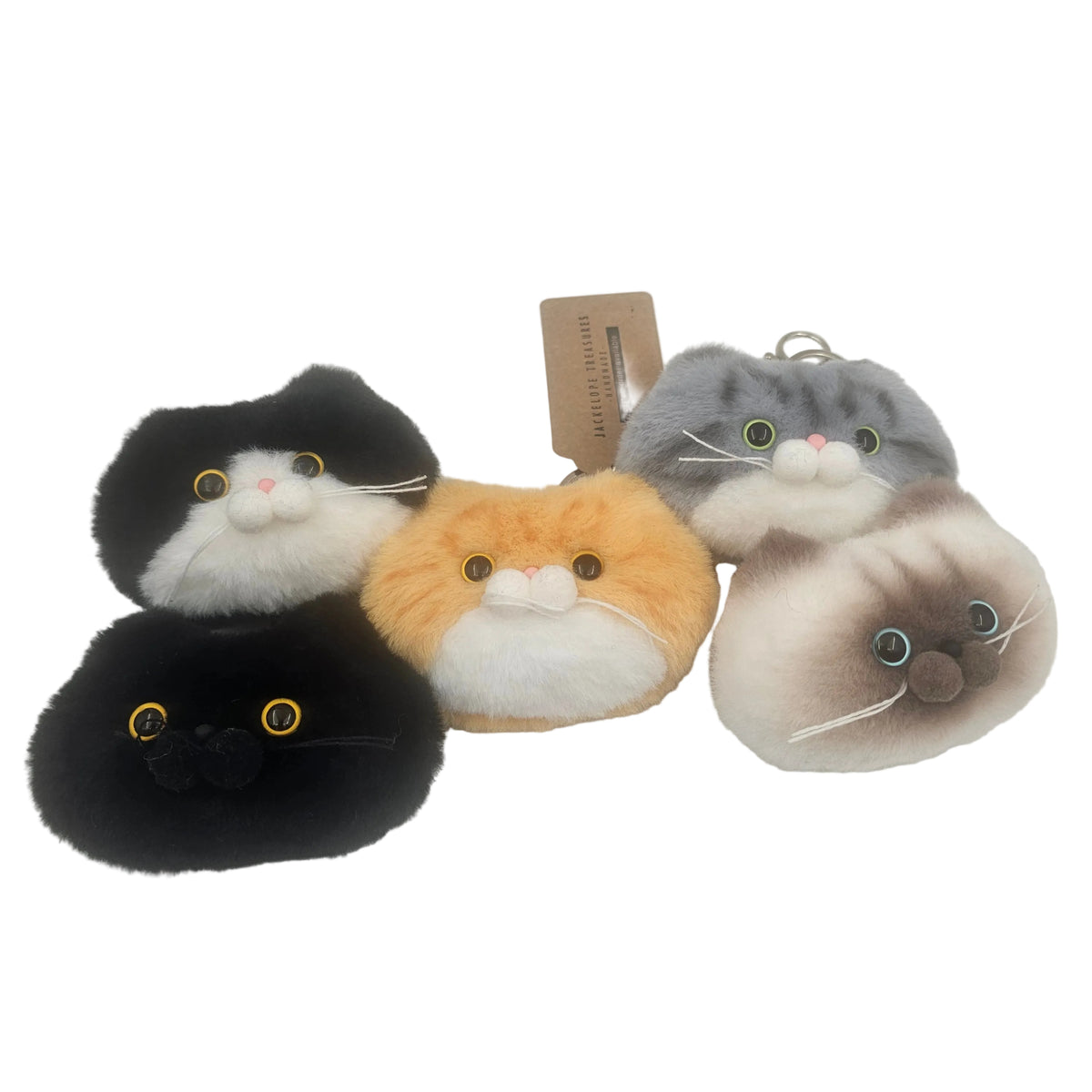 Four plush cat-face keychain bag charms  in different colors, including black and white, orange and white, gray and white, and cream and brown, arranged on a white background with a small paper tag in the center.
