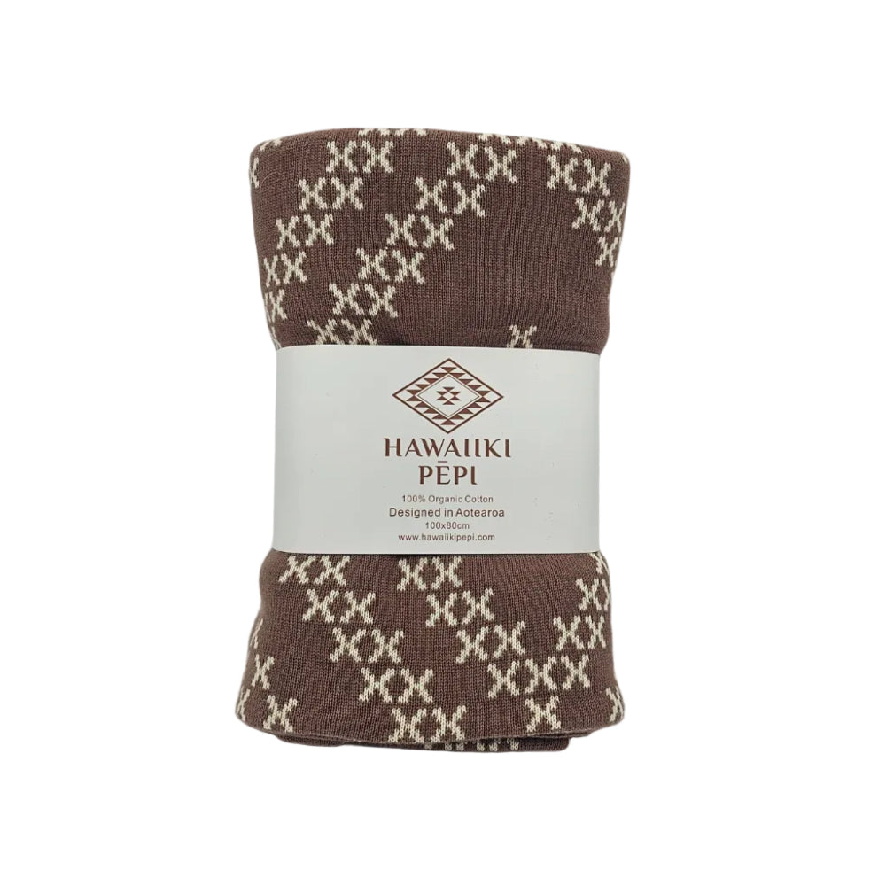 A rolled brown knit blanket with a cream geometric pattern, wrapped in a white paper band labeled 'HAWAIIKI PÉPI'.