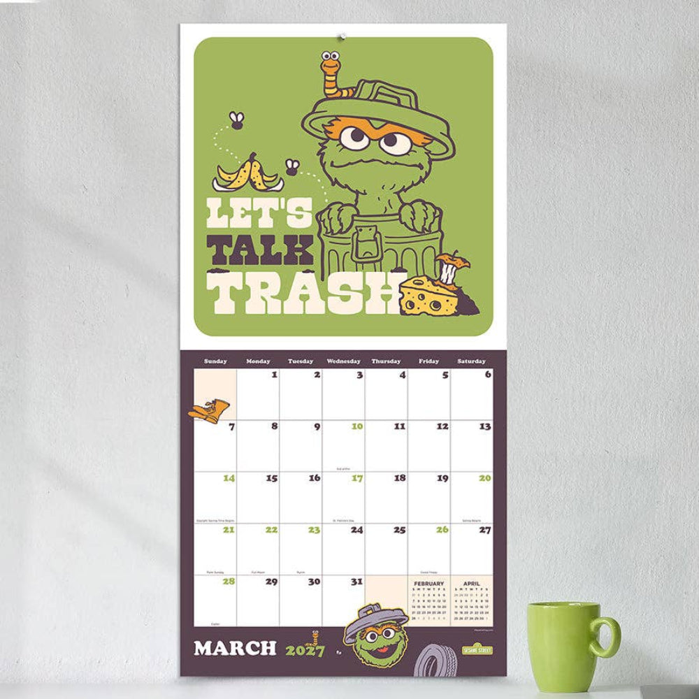 A March 2027 desk calendar featuring a green and purple Easter-themed design with a cartoon trash can character and the phrase 'LET'S TALK TRASH.' The calendar is shown standing upright against a light gray wall, with a green mug on a white surface below it.