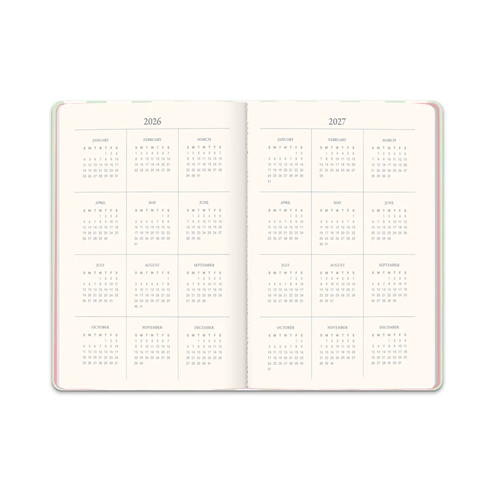 An open planner or notebook showing two interior calendar spreads for 2026 and 2027, with monthly grids on warm off-white pages and a thin multicolor border visible along the left and right edges.