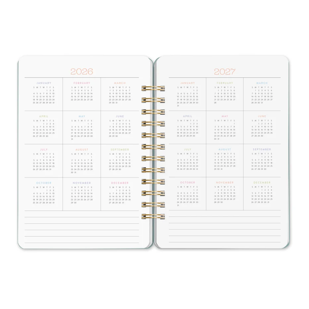 An open spiral-bound planner showing the pages for 2026 and 2027, with monthly calendars in a grid format and lined writing sections at the bottom. The pages are white with gold spiral binding and small pastel-colored month headers.