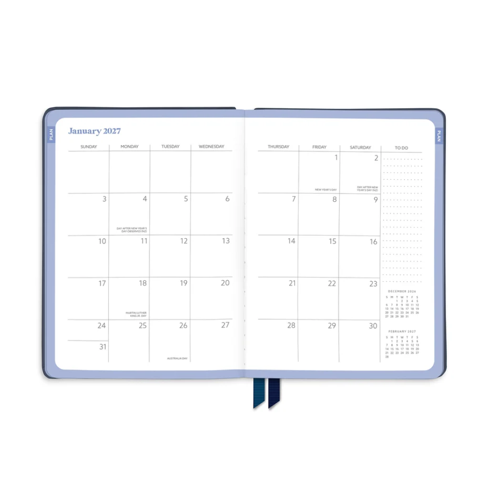 An open planner showing January 2027 with a light blue cover and navy ribbon bookmarks, photographed on a clean white background.
