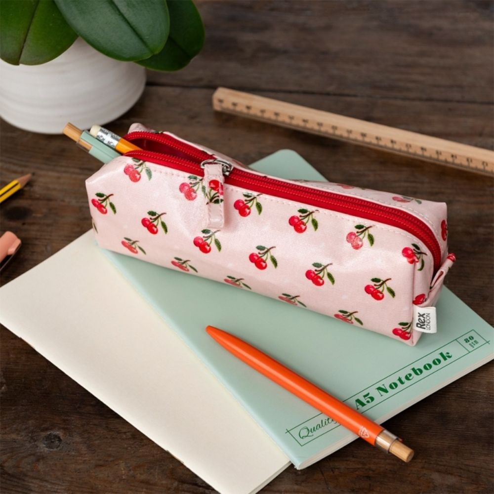 A pink zippered pencil case with a cherry print pattern, shown on a desk with a notebook, pencil, and ruler.