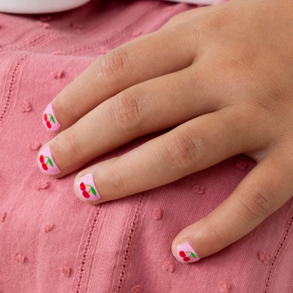 A close-up of a child's hand resting on a pink textured fabric, showcasing short fingernails decorated with a cherry pattern on a light pink background.
