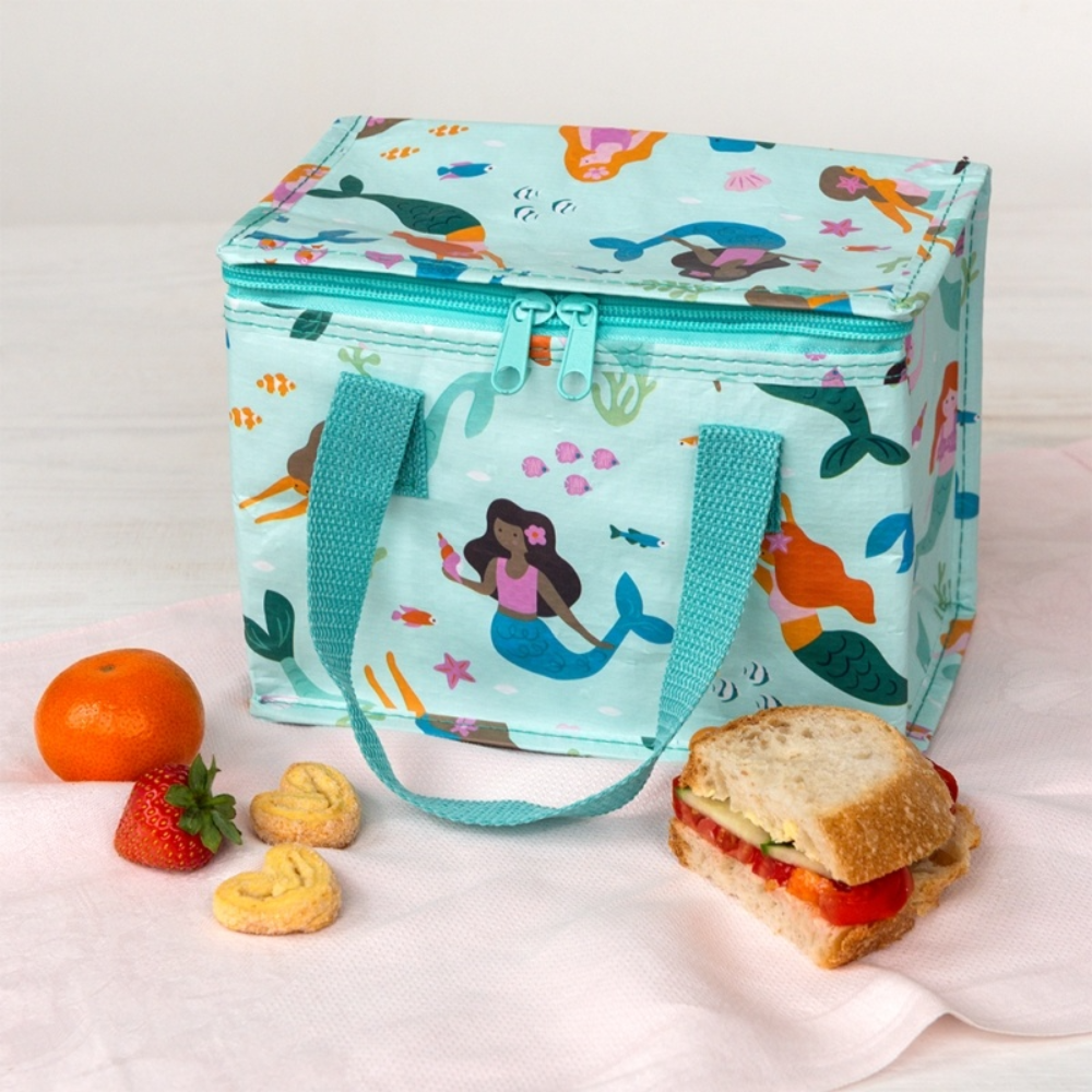 A light aqua insulated lunch bag with an underwater mermaid print, displayed on a white cloth with a sandwich, a strawberry, an orange, and cookies around it.