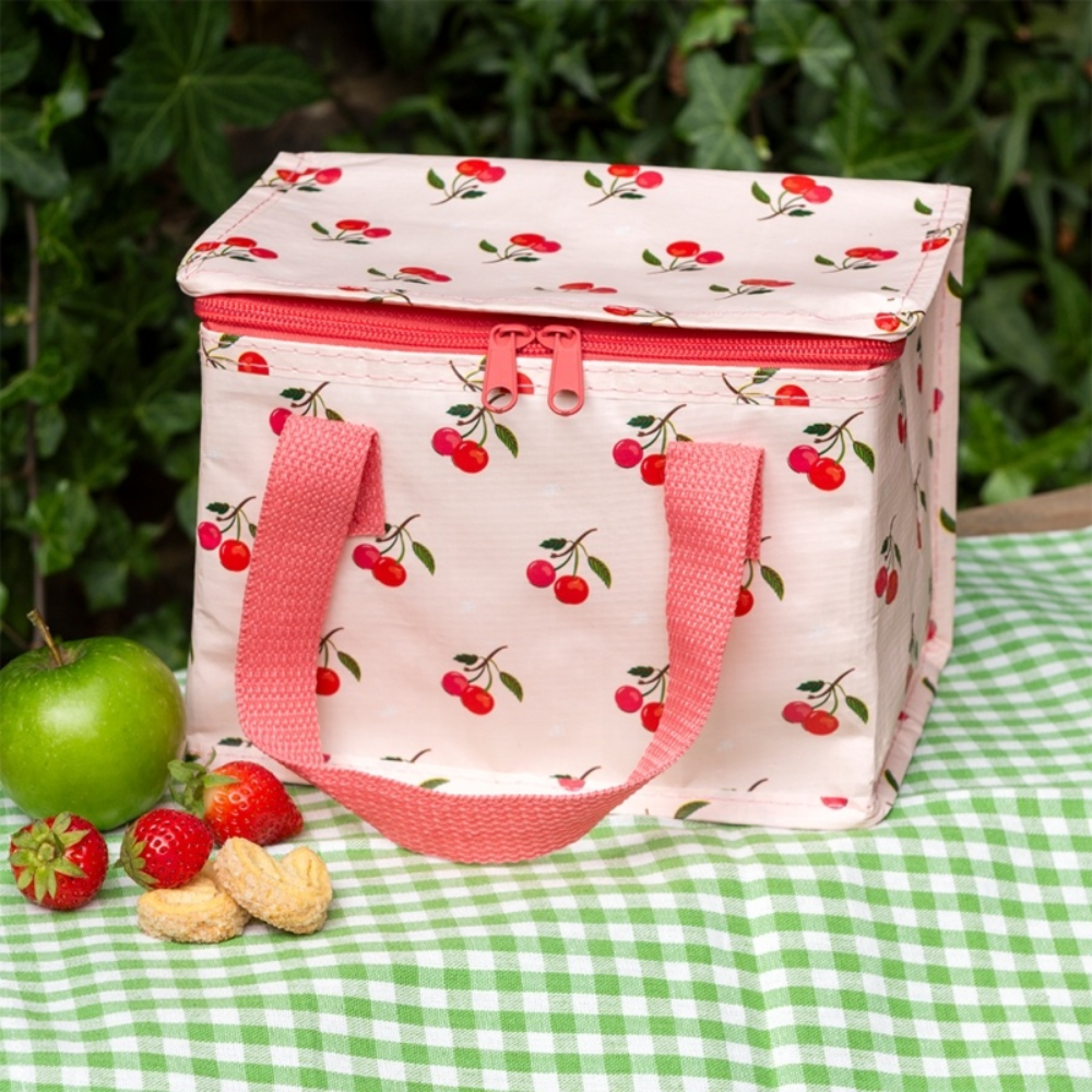 A cherry-patterned insulated lunch bag with pink handles and a pink zipper, placed on a green-and-white checkered blanket outdoors with a green apple, strawberries, and peanuts nearby.