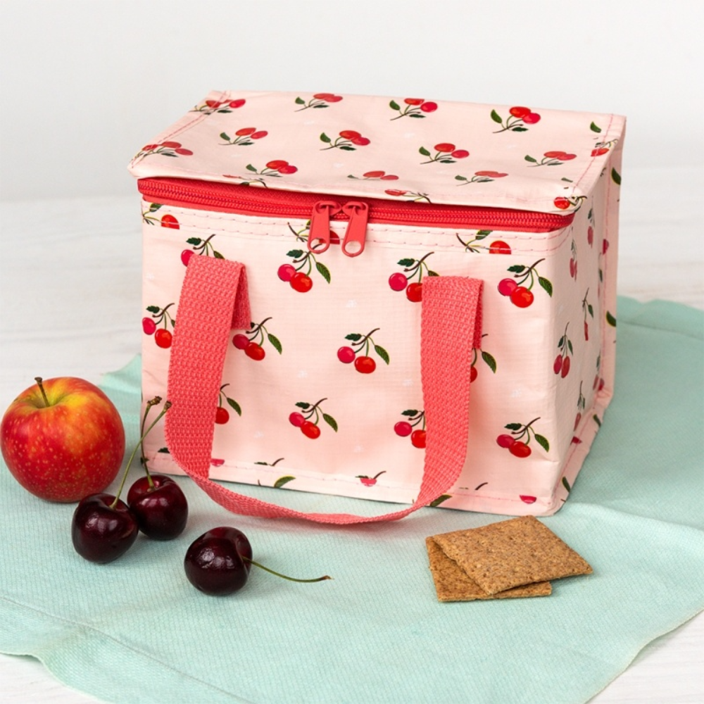 A pink fabric lunch bag with a cherry print pattern, red zipper, and red handles, styled on a mint green cloth with an apple, cherries, and crackers.