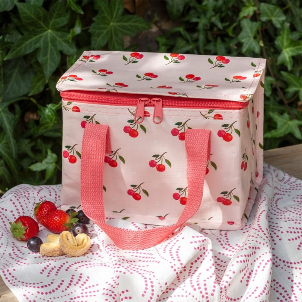 A pink insulated lunch bag with a cherry print and coral handles, set on a white picnic blanket outdoors with strawberries and crackers nearby.
