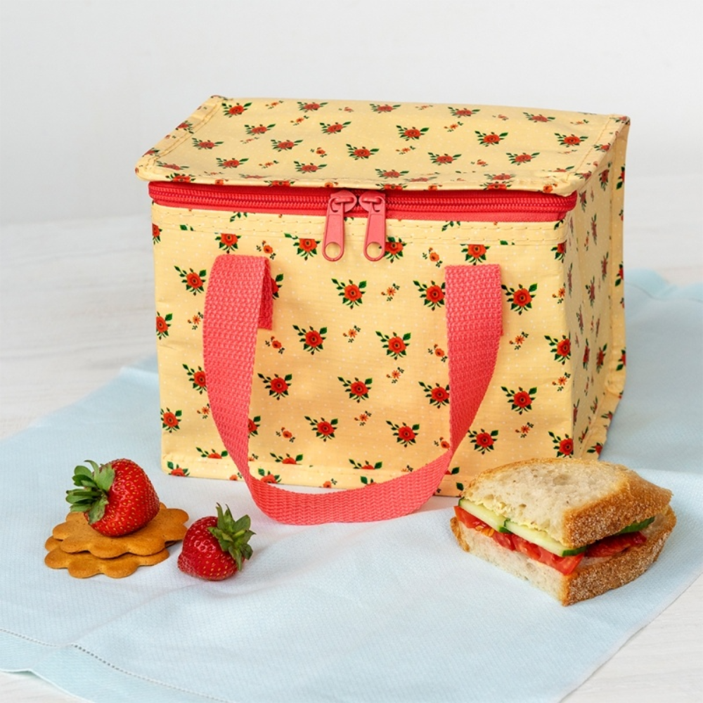 Floral-patterned lunch bag with pink handles on a light blue cloth, accompanied by strawberries and a sandwich.