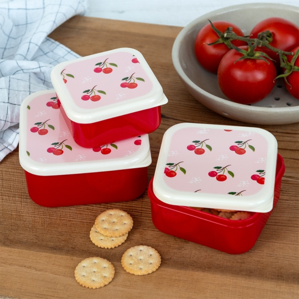 Set of red food containers with cherry design lids on a wooden surface with crackers and tomatoes.