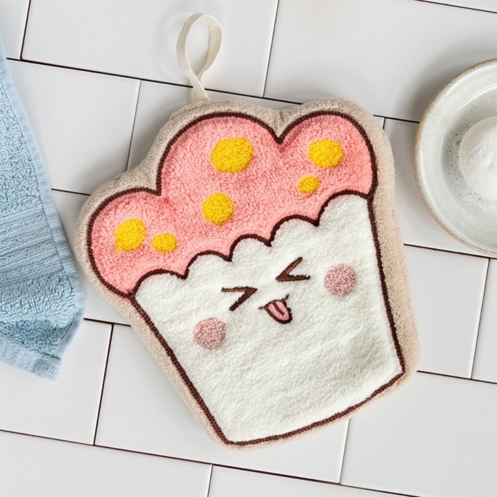 A plush cupcake-shaped kitchen towel or potholder with a cute cartoon face, featuring pink frosting with yellow polka dots and a white cake base, styled on a white tiled surface.