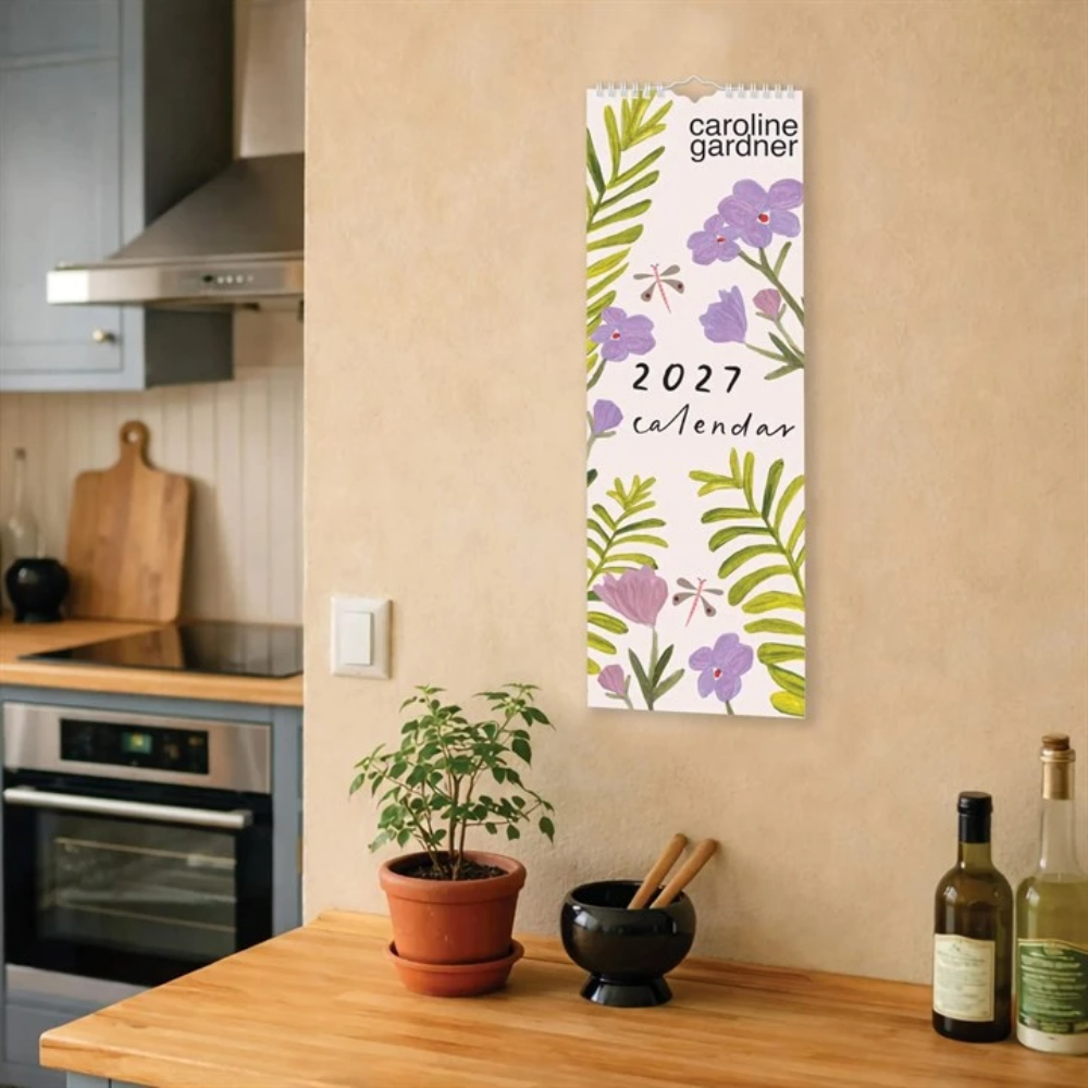 A vertical 2027 Caroline Gardner calendar with a botanical floral pattern hangs on a beige wall in a modern kitchen setting.