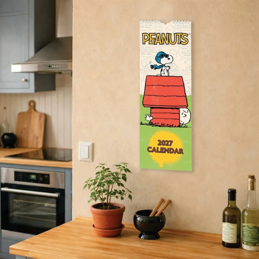 A 2027 Peanuts calendar hanging on a beige wall in a kitchen setting, featuring Snoopy and Woodstock on the doghouse with the text 'PEANUTS' at the top and '2027 CALENDAR' at the bottom.