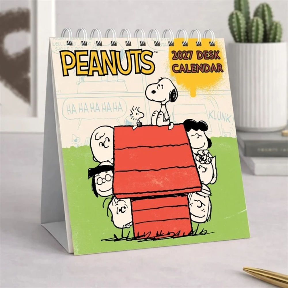 A 2027 Peanuts desk calendar standing on a white surface, featuring the Peanuts characters around a red doghouse on a green background. The calendar has a spiral wire binding at the top and is photographed in a clean indoor setting with a potted cactus blurred in the background.