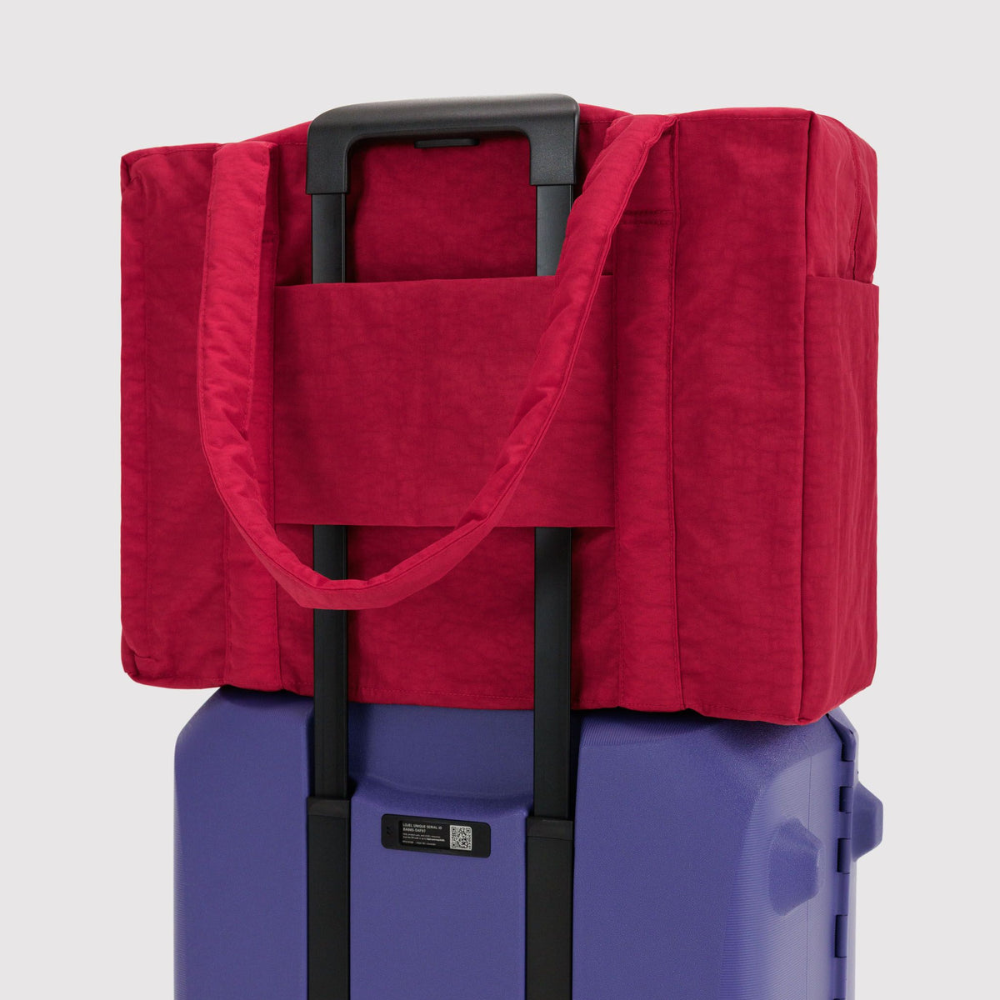 A red soft-sided rolling duffel bag with black handles, shown resting on top of a purple rolling suitcase against a light gray background.