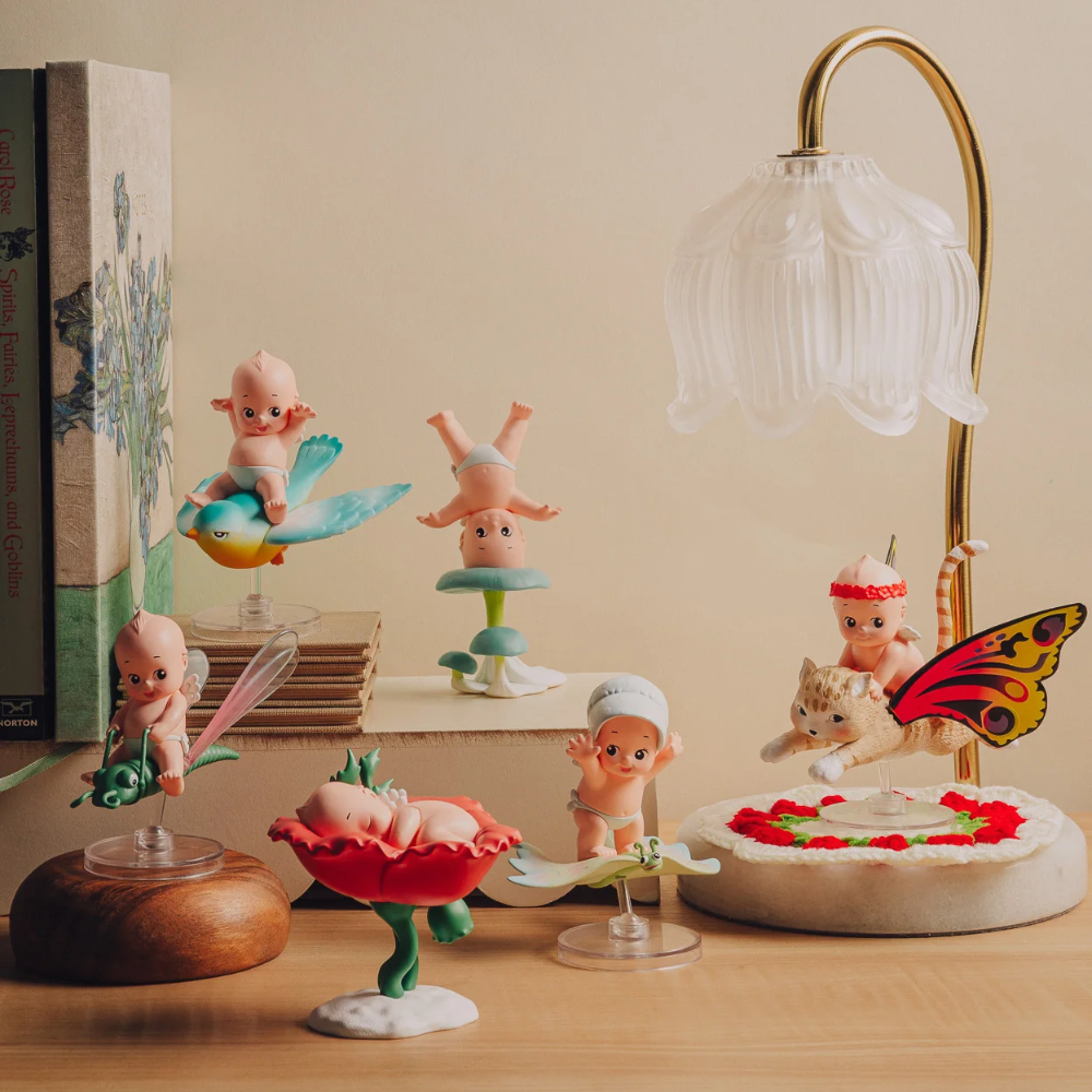 A styled tabletop display of small decorative figurines and objects, including pastel cherub-like dolls, a translucent bell-shaped glass lamp, a butterfly decoration, a flower-shaped dish, and books, arranged on a wooden surface against a warm beige wall.