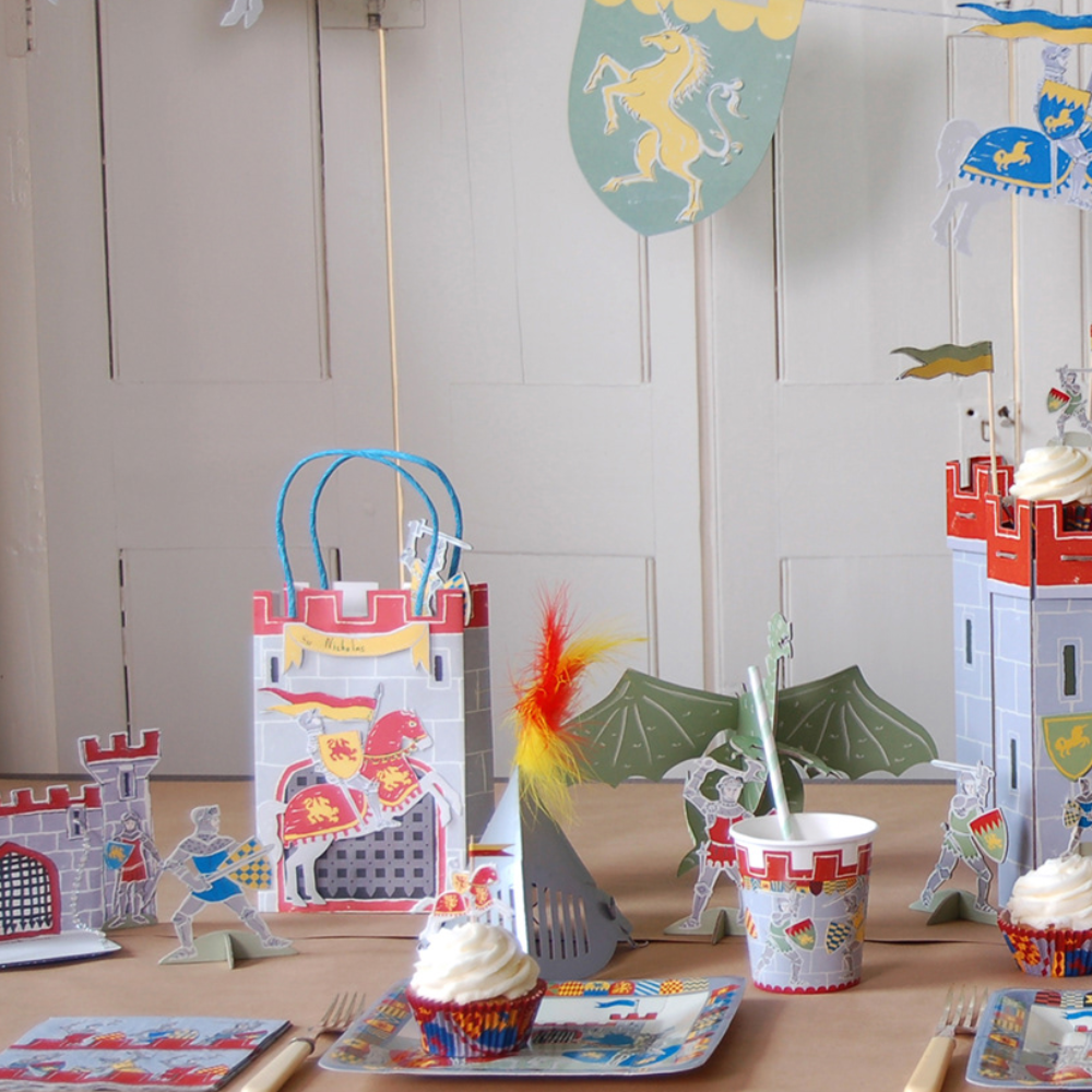 A medieval knight and castle themed party table setting featuring a large castle centerpiece, paper banners, cups, plates, napkins, and gift bags decorated with knights, dragons, and heraldic shields.