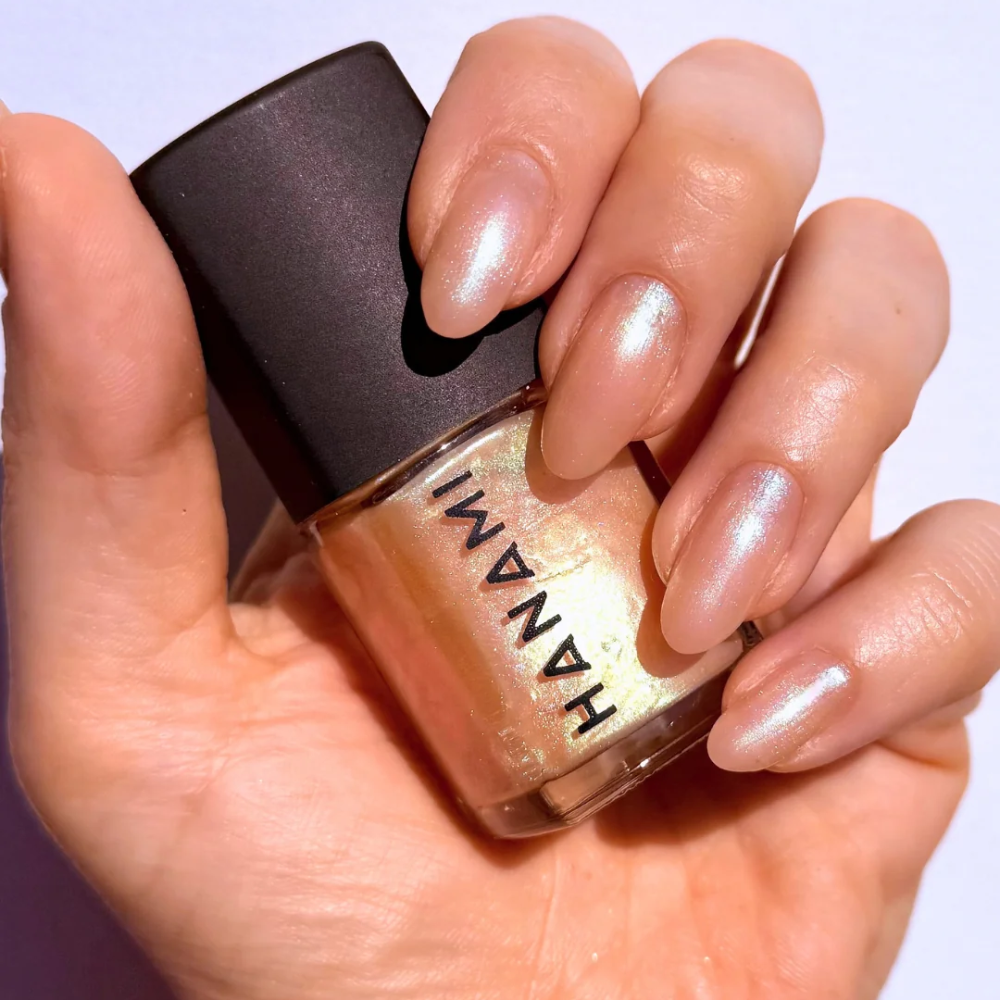 A close-up of a hand holding a bottle of Hanami nail polish. The nails are painted with a shimmering, metallic champagne-gold polish, and the bottle cap is black.