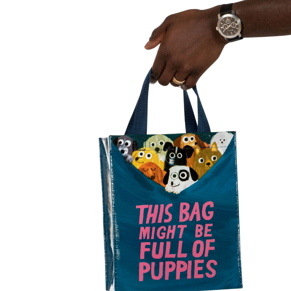 A dark teal reusable tote bag with pink text reading 'THIS BAG MIGHT BE FULL OF PUPPIES' and a window showing colorful cartoon puppies peeking out, held by a person's hand against a white background.