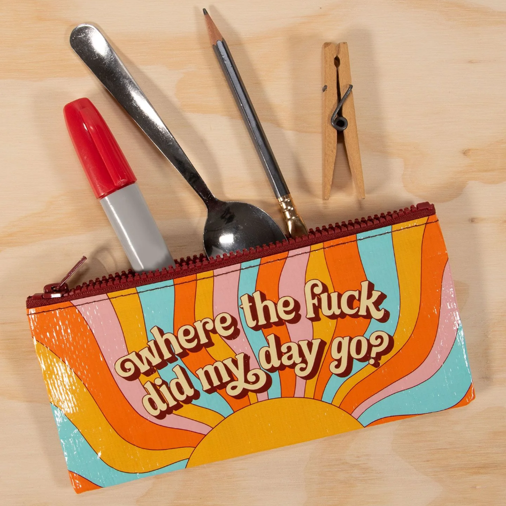 A colorful zippered pouch with a retro sunburst design and the text 'where the fuck did my day go?' lying on a light wood surface, with a lipstick, spoon, pencil, and wooden clothespin protruding from the top.