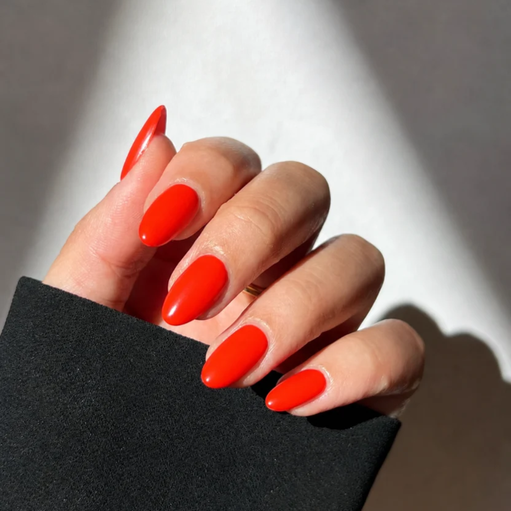 A close-up of a hand with long, almond-shaped nails painted in a bright, glossy neon orange-red color, wearing a black sleeve against a light gray background with dramatic shadows.