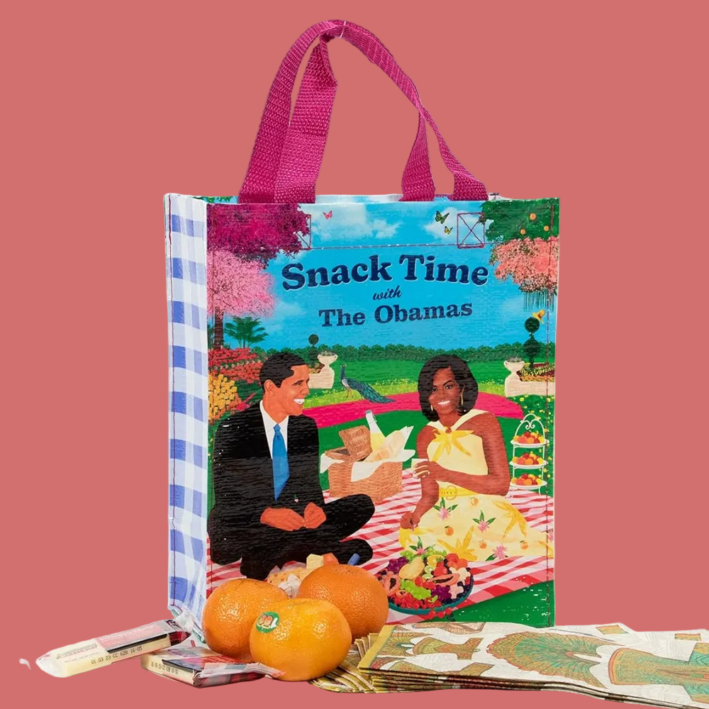 A reusable tote bag with pink handles stands on a white studio background. The front of the bag features an illustrated picnic scene with the text 'Snack Time and The Obamas.