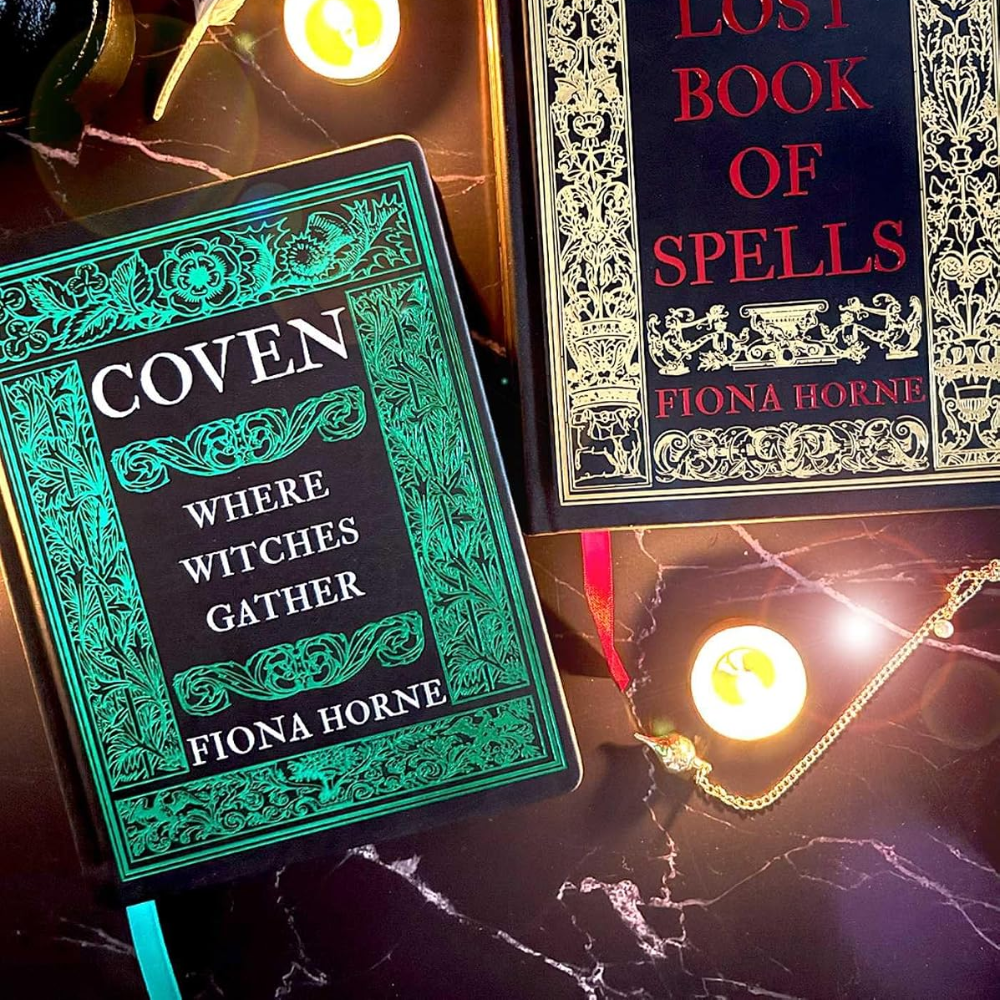 Two hardcover books by Fiona Horne, 'Coven: Where Witches Gather' and 'Lost Book of Spells', displayed on a dark marble surface with mystical props including a glowing candle, a feather quill, and a gold chain.