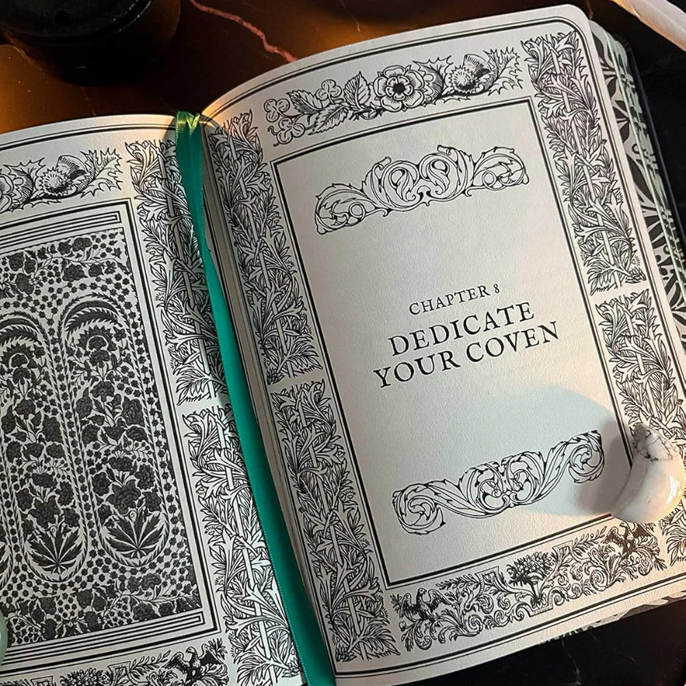 An open decorative book or journal with ornate black floral border illustrations on both pages. The right page reads 'CHAPTER 1 DEDICATE YOUR COVEN' in serif text, and a teal ribbon bookmark hangs from the center.