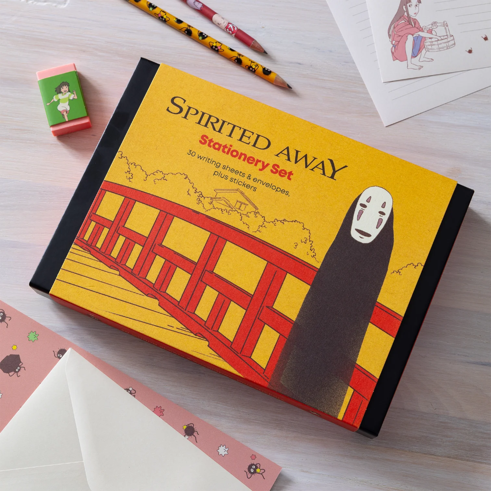 A boxed stationery set titled 'Spirited Away' lies on a light wood desk, surrounded by stationery props including pencils, a small green eraser, a pink patterned card, and white envelopes. The box art shows a yellow background, a red train, and the character No-Face.