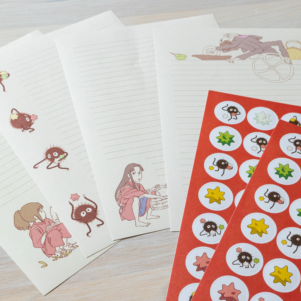 A set of stationery items laid out on a light wood surface, including cream lined and blank memo pads with Sanjiro and Neemon character illustrations, and a red sheet of round stickers featuring the same characters and star shapes.