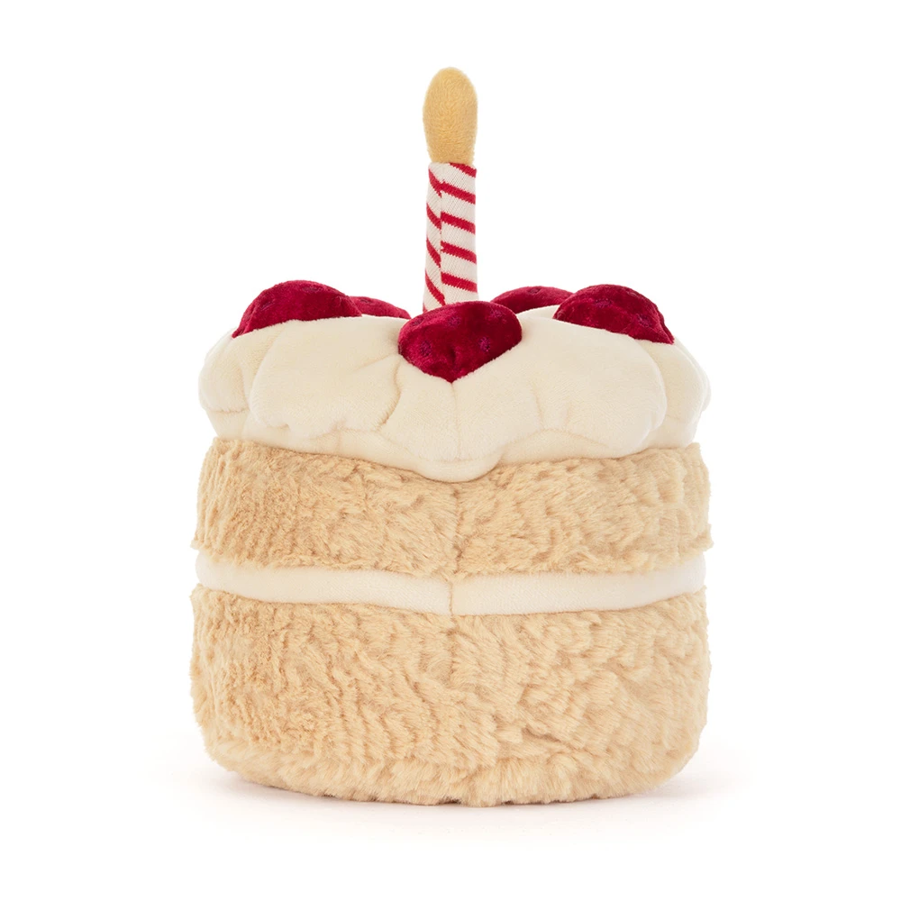 A plush toy shaped like a birthday cake, featuring a textured beige body, cream-colored frosting, red velvet-like frosting on top, and a single red-and-white striped candle with a yellow tip, set against a plain white background.