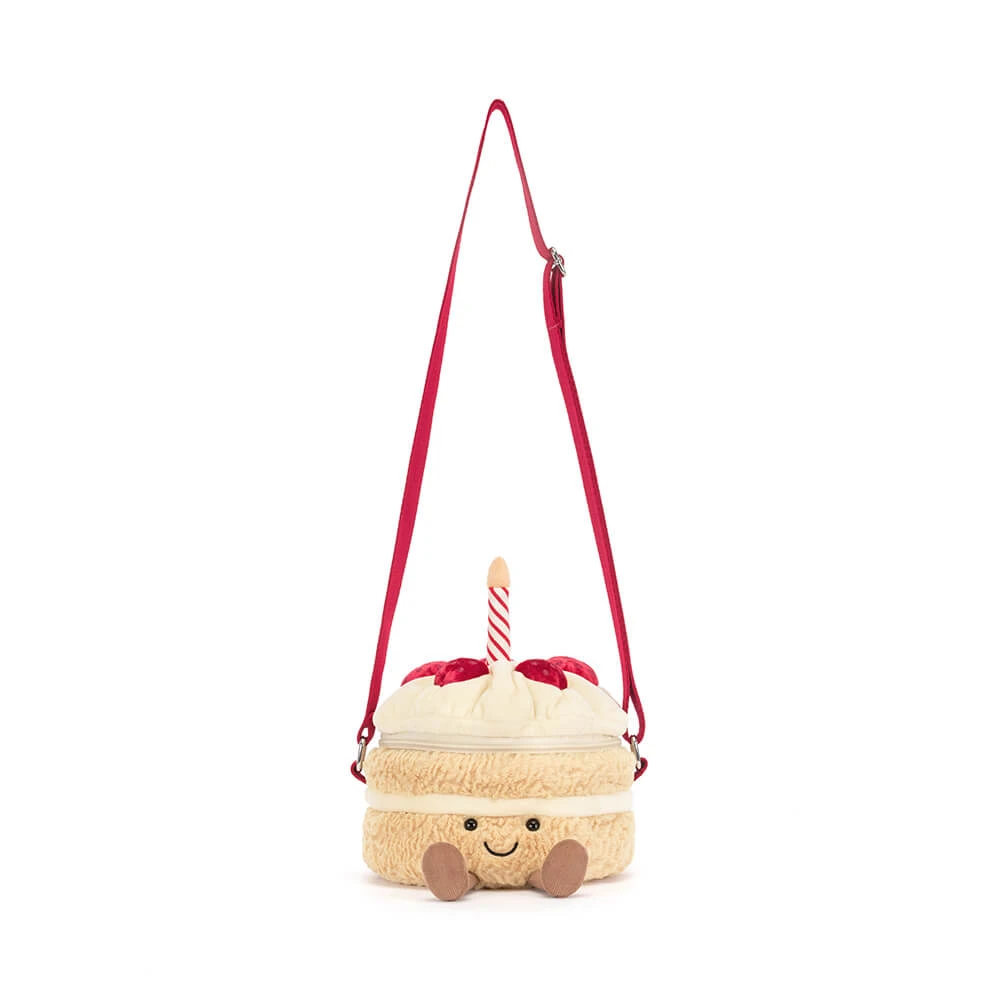 A plush cake-shaped shoulder bag with a cream-colored fuzzy body, red frosting details, a red and white striped candle, and a red adjustable strap, shown on a plain white background.