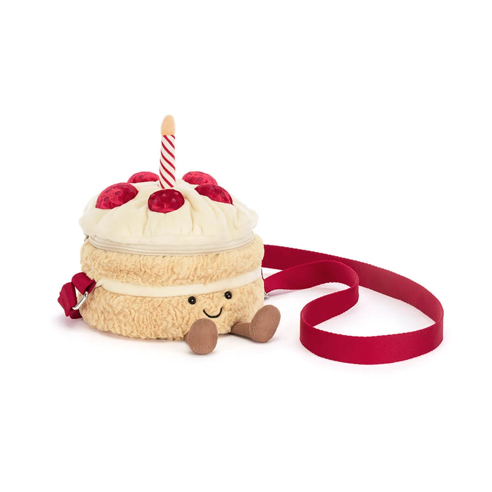 A plush cake-shaped crossbody bag with a red ribbon strap, featuring cream frosting, red cherries, a candle, and a smiling face on a white background.