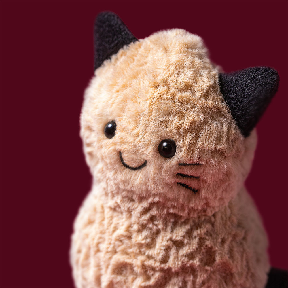A close-up of a plush cat toy with fluffy cream-colored fur, black ears, and a simple black embroidered smiley face, set against a solid dark red background.
