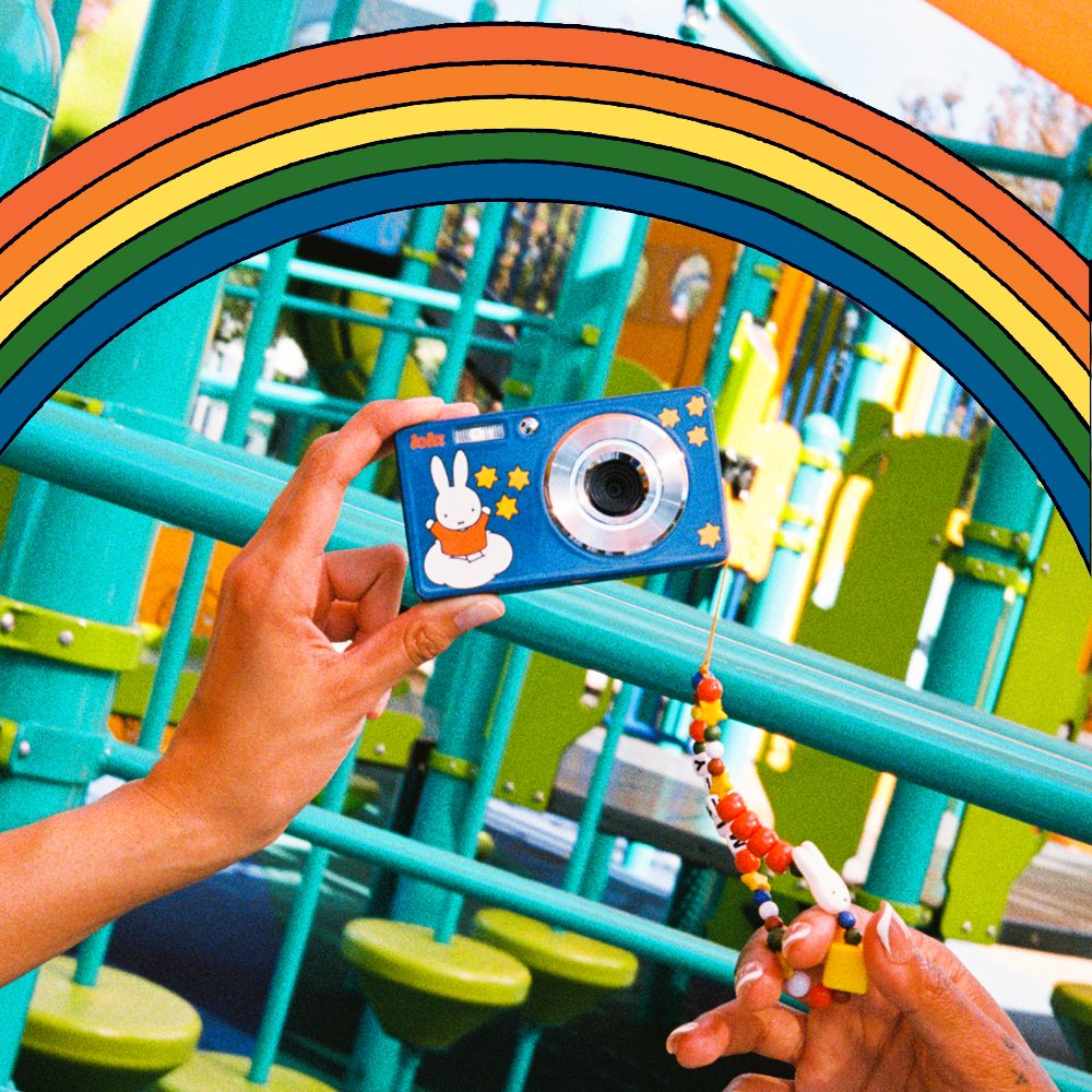 A blue compact camera with a bunny illustration on the front is held by two hands in front of a colorful playground with a rainbow graphic overlay. A beaded wrist strap is also held in the right hand.