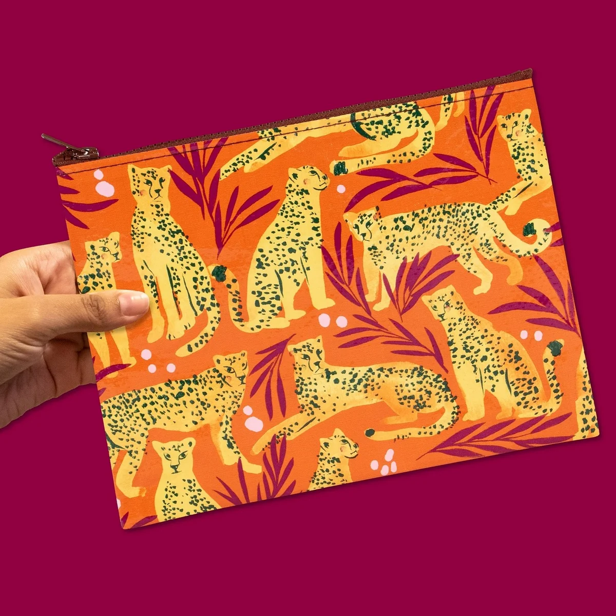 A hand holding a zippered pouch with a bold orange background and an allover print of yellow leopards and dark green foliage, set against a solid magenta background.