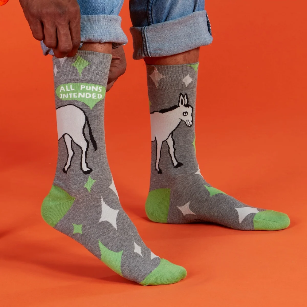 Gray crew socks with green and white star patterns, a donkey illustration, and the text 'ALL PUNS INTENDED' on the side, shown on a person's feet against an orange background.