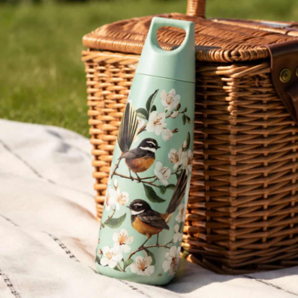 A mint green insulated water bottle with a printed design of two birds and white blossoms, leaning against a wicker picnic basket on a white blanket with grass in the background.